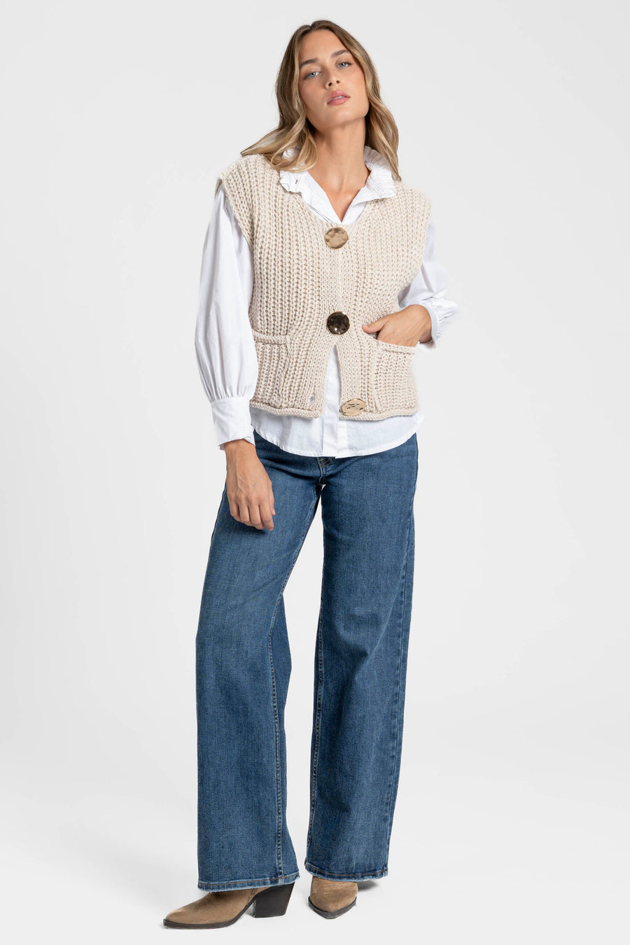 Femme portant un gilet sans manches en maille femme crème avec boutons métalliques, avec un pantalon en denim pour un look chic. Gilet maille femme.