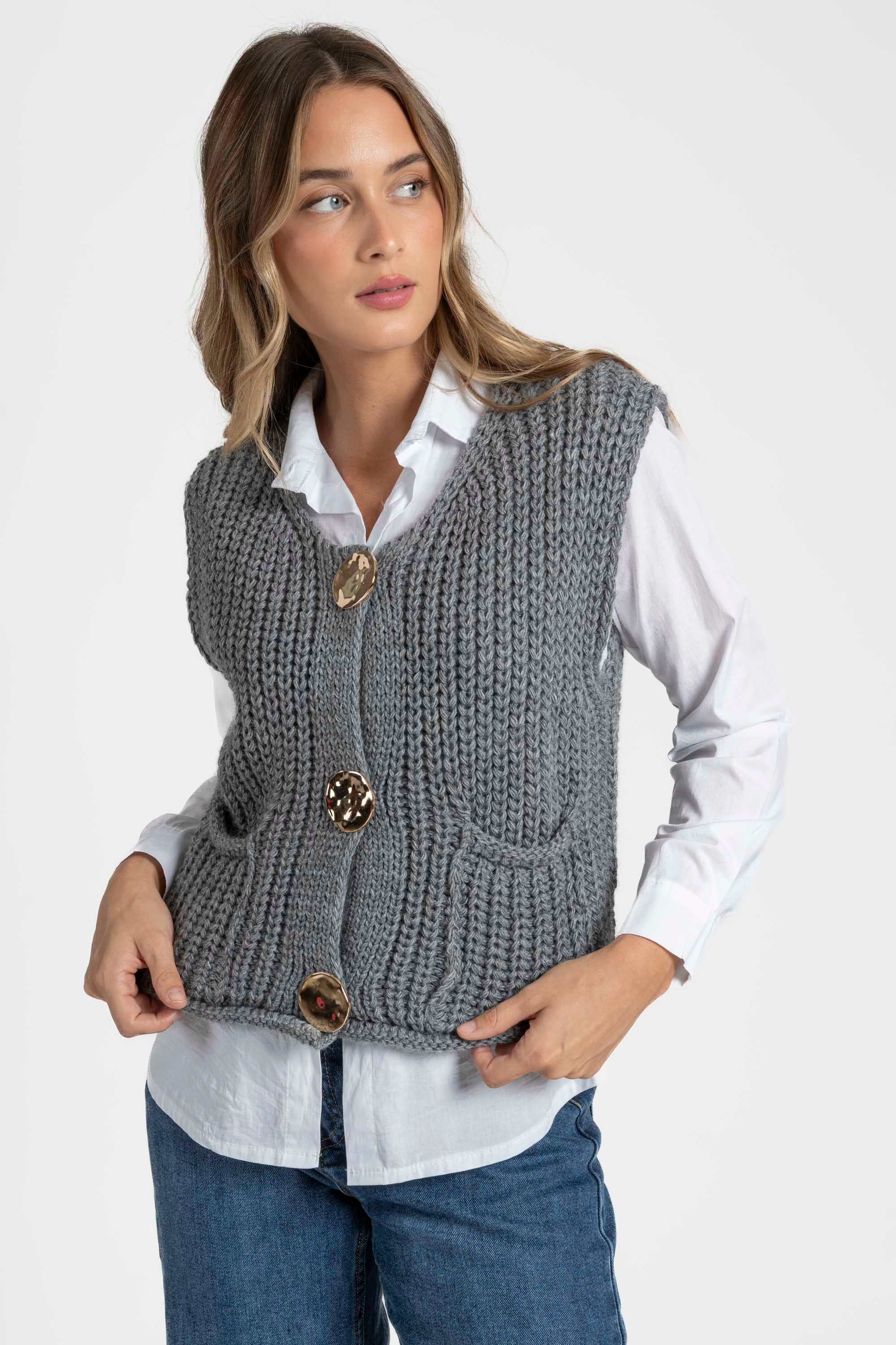 Gilet sans manches en maille femme de couleur grise présenté avec une chemise blanche, boutonnière métallique et poches avant pratiques. Gilet maille femme.