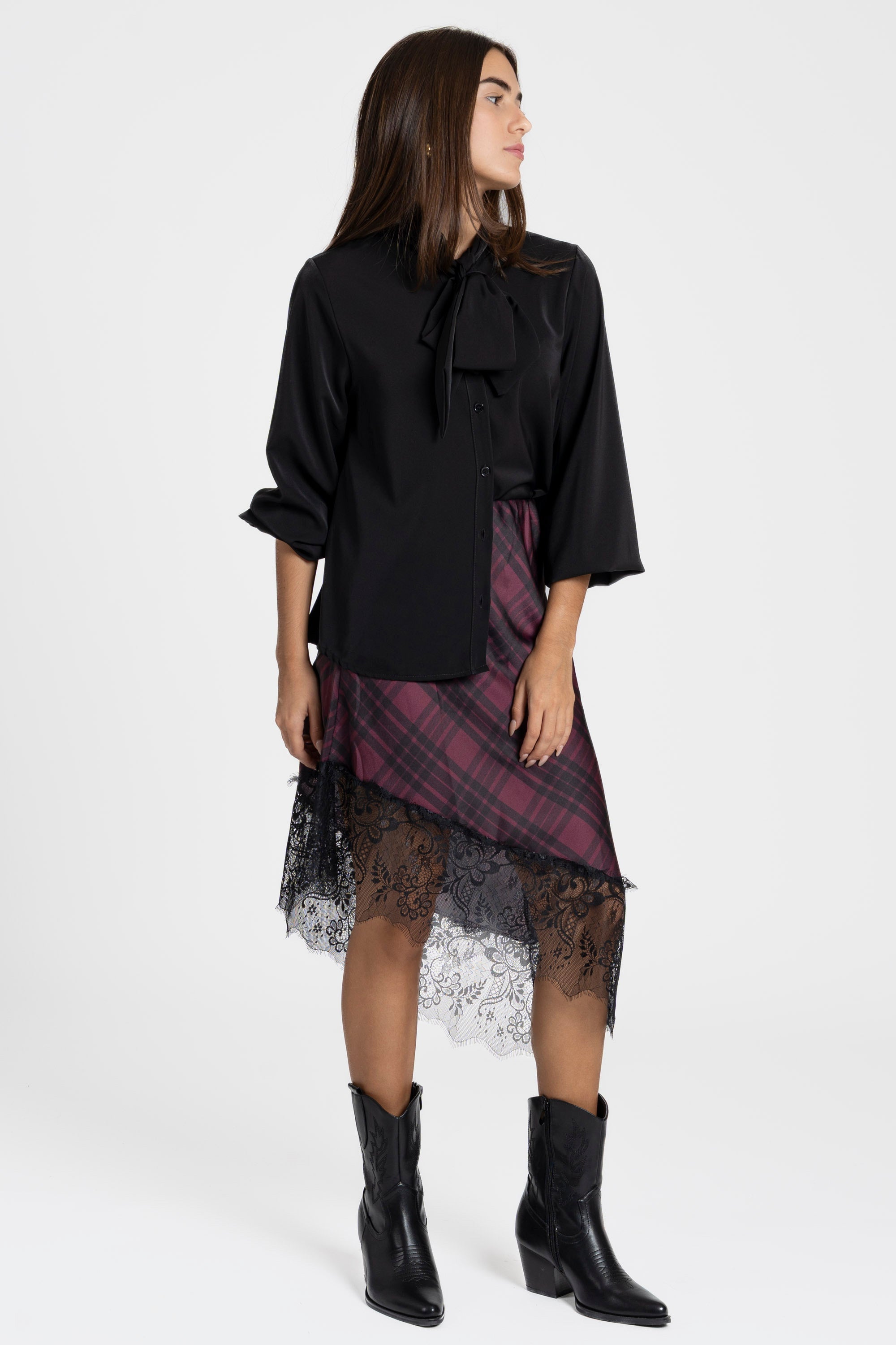 Femme avec une silhouette élégante portant une jupe midi satinée asymétrique en satin, se tenant debout avec des bottes noires.