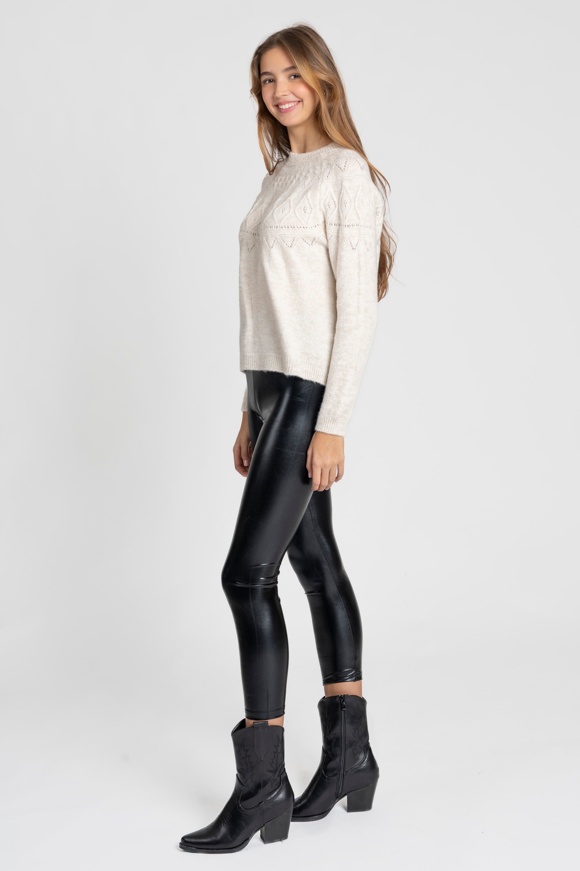 Vue latérale d'une femme en legging noir brillant taille haute, coupe ajustée, avec un pull clair et des bottes noires, apportant une touche moderne.