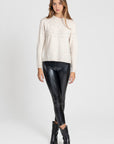 Femme en legging noir brillant taille haute, coupe ajustée, posant avec un pull beige en tricot et des bottes noir, pour un style élégant.