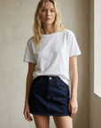 Femme se tenant dans une belle lumière naturelle, portant une mini jupe en jean avec un t-shirt blanc.