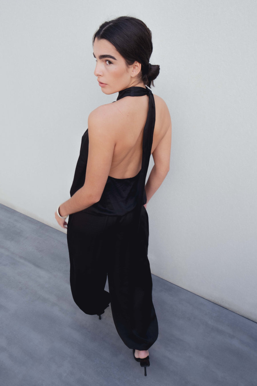 Vue de dos d'une femme en pantalon bombacho satiné noir, jetant un regard vers la gauche, mettant en avant la coupe élégante de son haut.