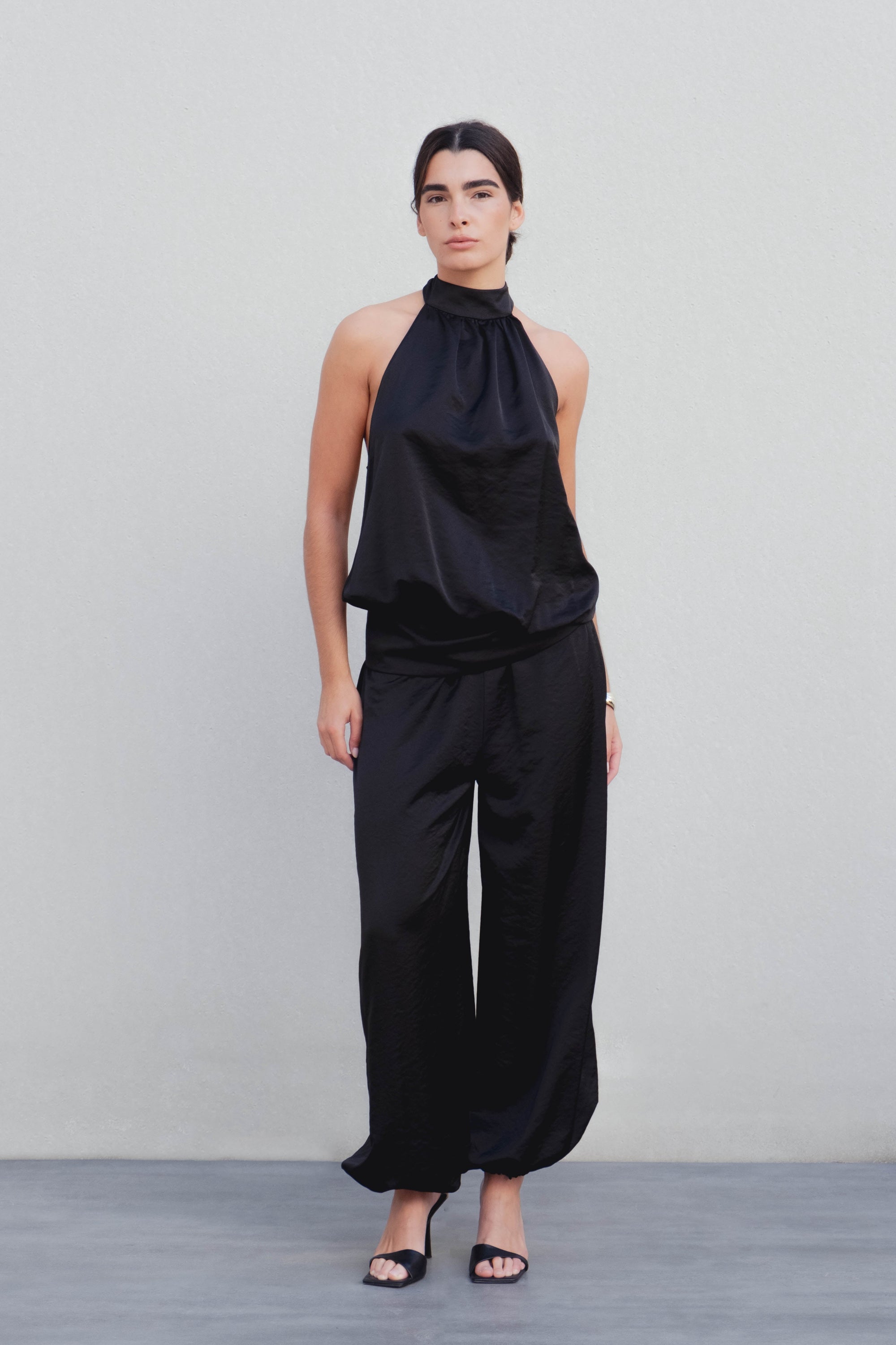 Femme portant un pantalon bombacho satiné noir avec un haut assorti, se tenant debout sur un fond neutre. Idéal pour des soirées chic.