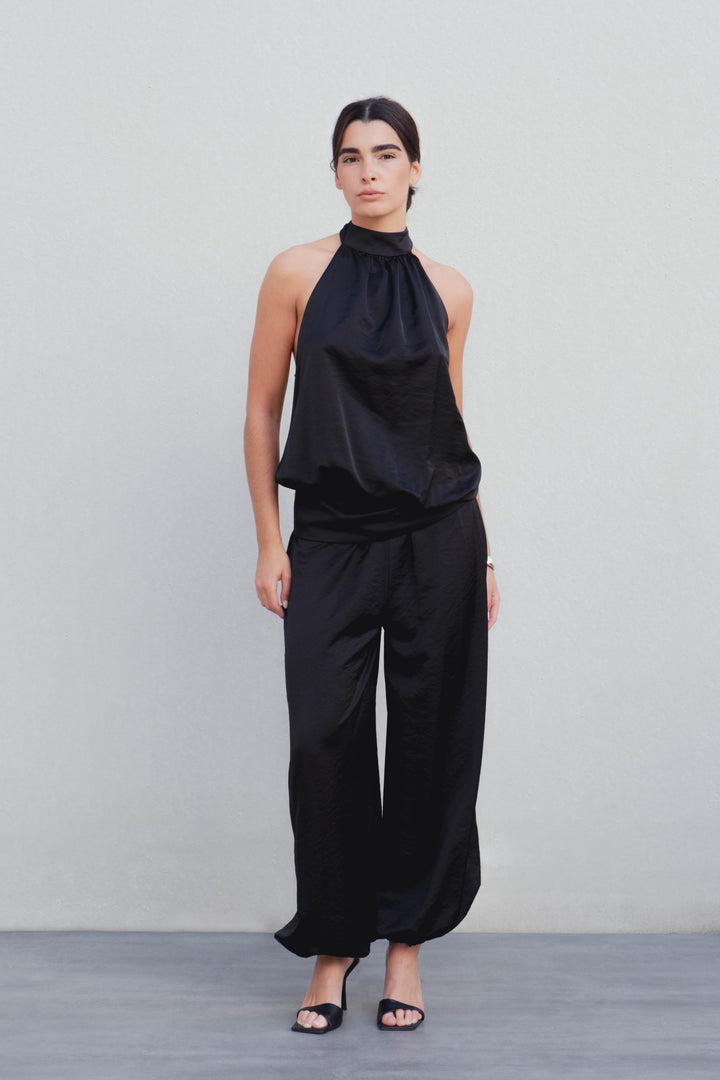 Femme portant un pantalon bombacho satiné noir avec un haut assorti, se tenant debout sur un fond neutre. Idéal pour des soirées chic.