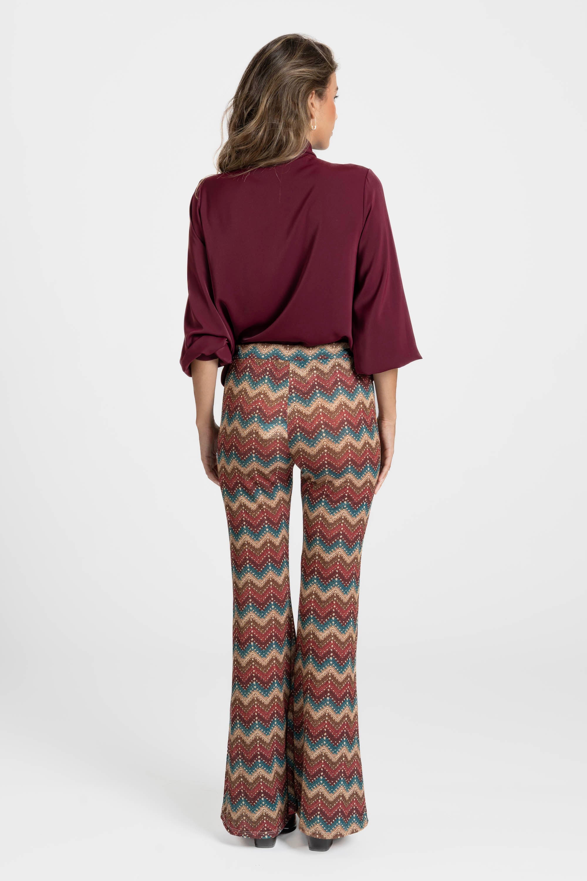 Femme montrant le dos avec un pantalon flare taille haute fluide imprimé en zigzag multicolore et une blouse bordeaux.