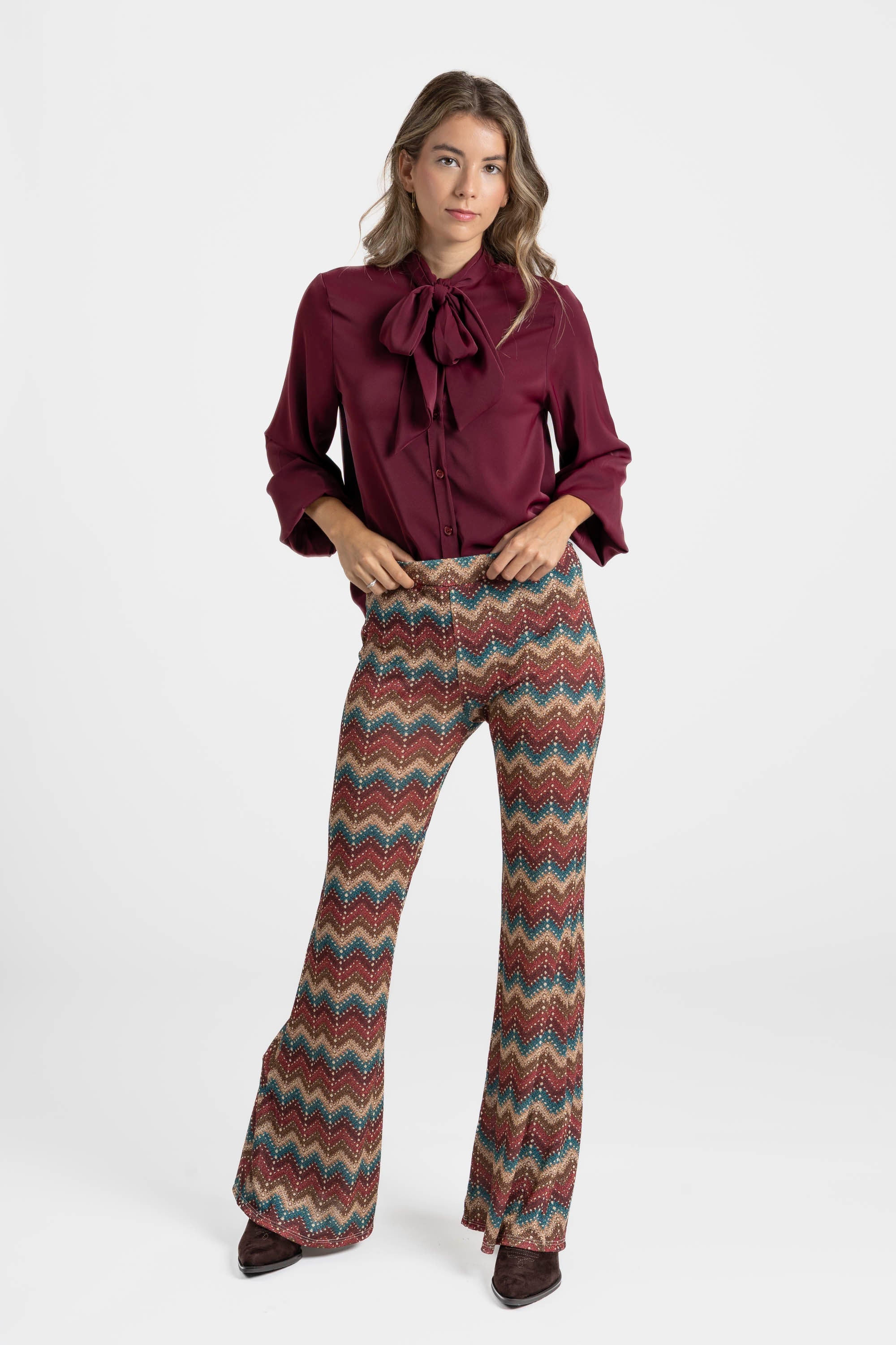 Femme souriante portant un pantalon flare taille haute fluide imprimé en zigzag multicolore et une blouse bordeaux.