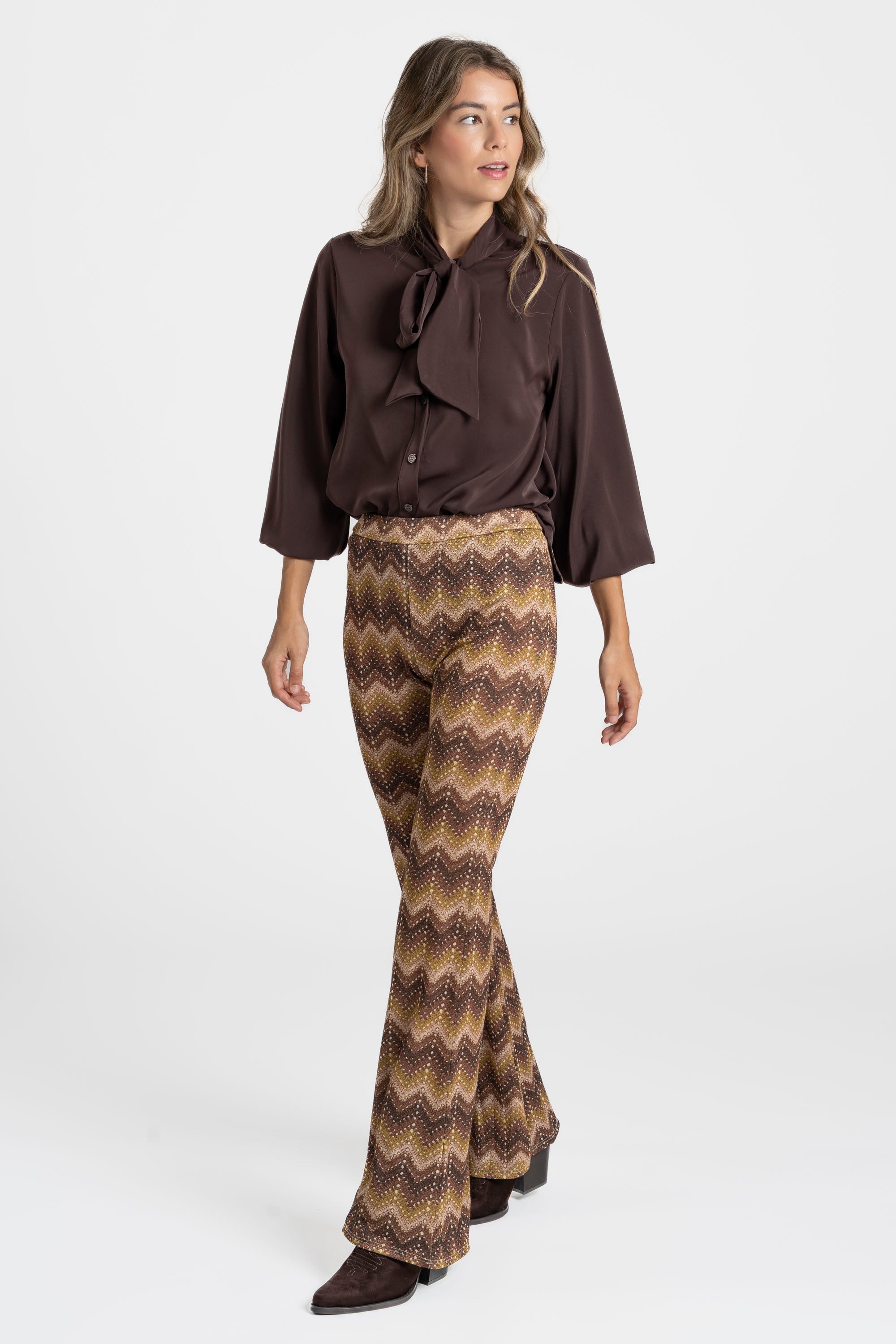 Femme élégante portant un pantalon flare taille haute fluide imprimé en zigzag marron, accompagnée d'une blouse marron.