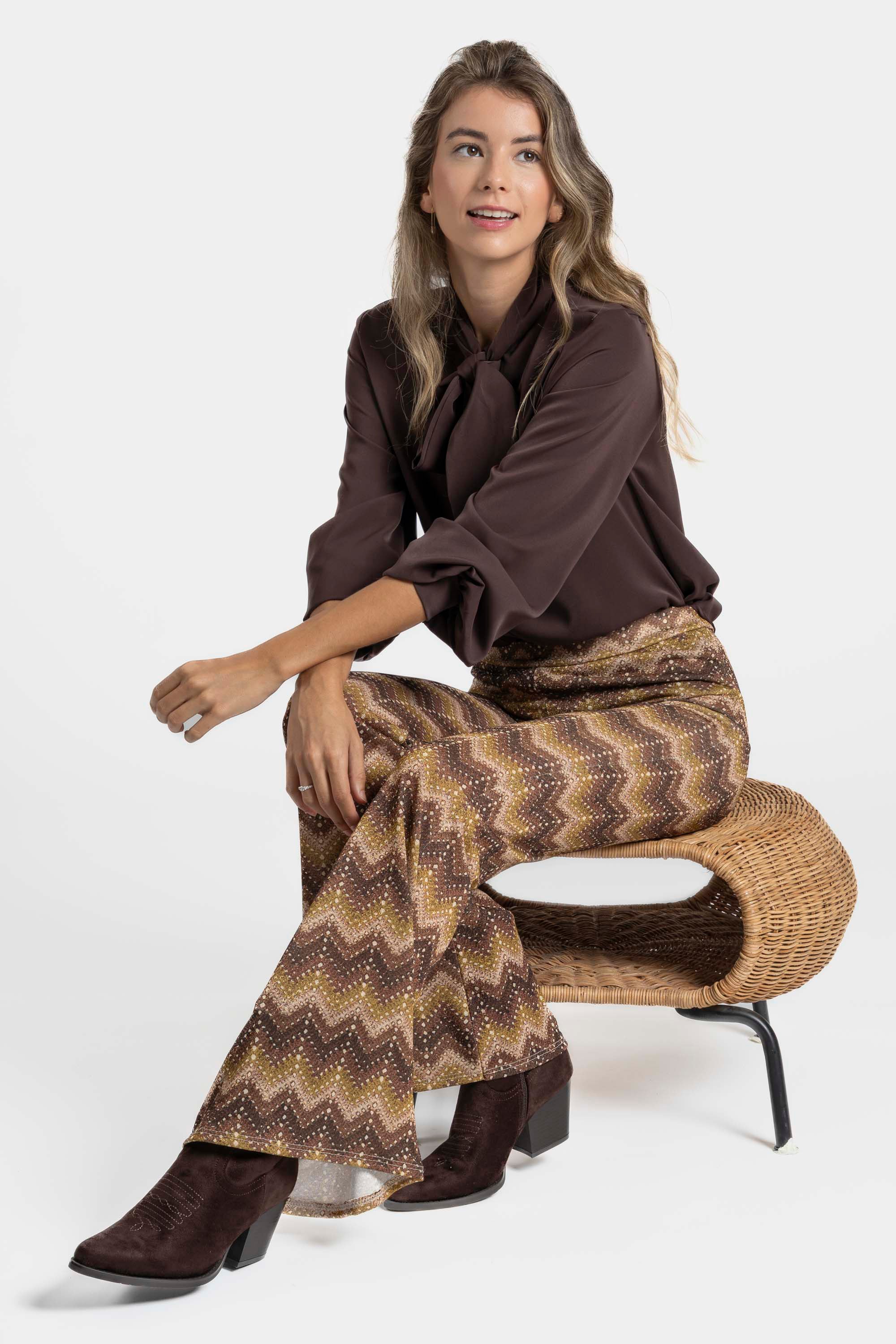 Femme assise sur un tabouret, affichant un pantalon flare taille haute fluide imprimé en zigzag marron, accompagnée d'un haut marron.