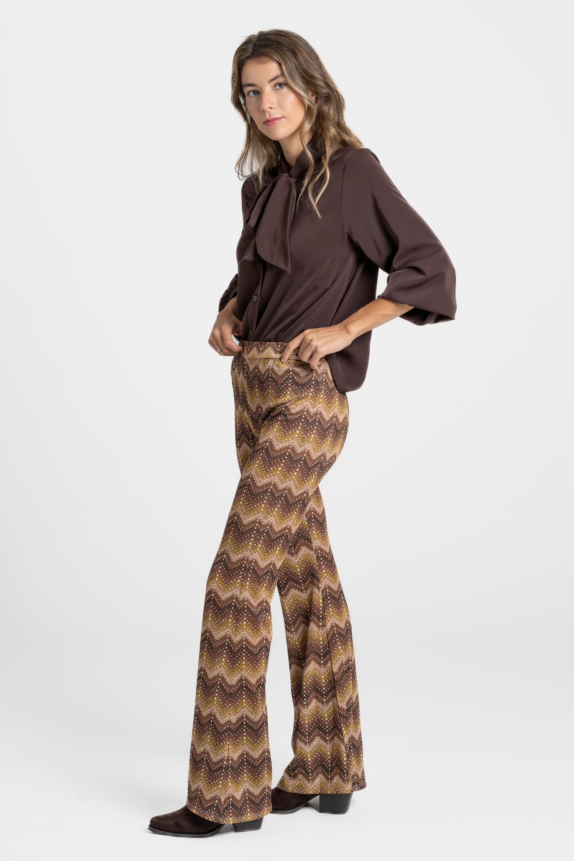 Femme debout en tenue chic, portant un pantalon flare taille haute fluide imprimé en zigzag marron avec une blouse marron.