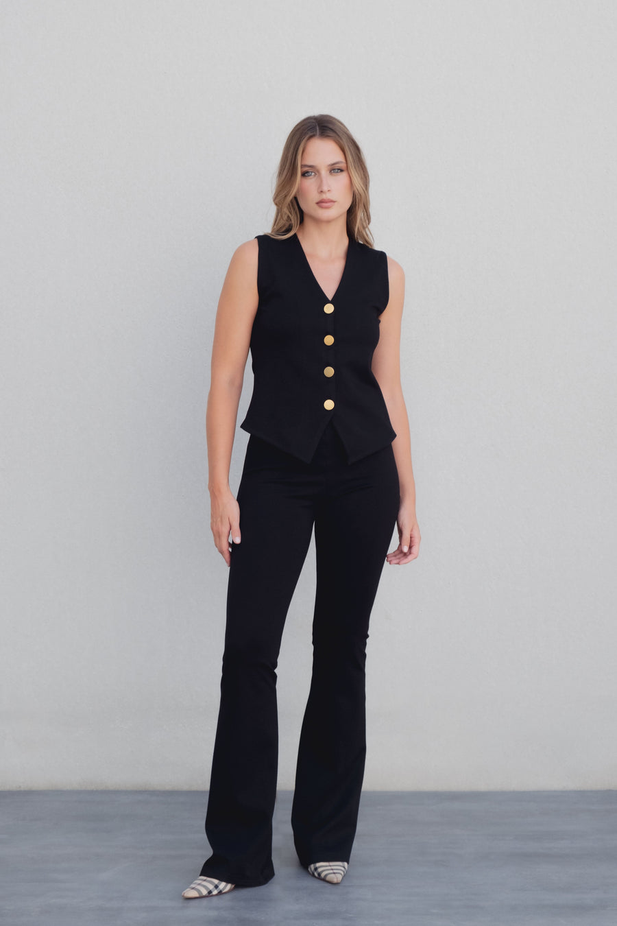Femme debout dans un pantalon flare taille haute noir texturé en viscose extensible, mettant en valeur sa silhouette avec un haut noir à boutons dorés.