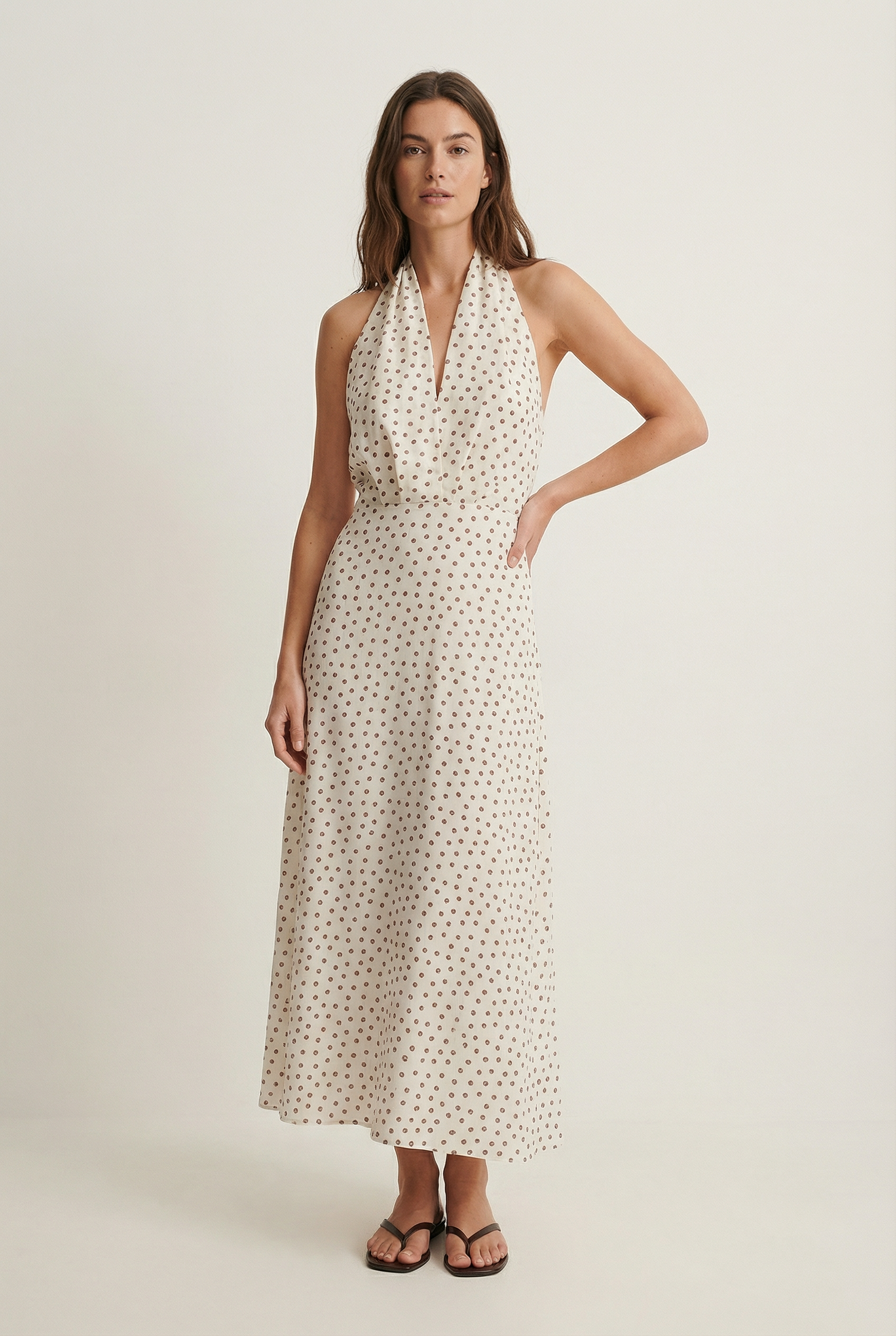 Femme debout dans une robe midi imprimée écru à pois, col halter décolleté plongeant, sur fond neutre.
