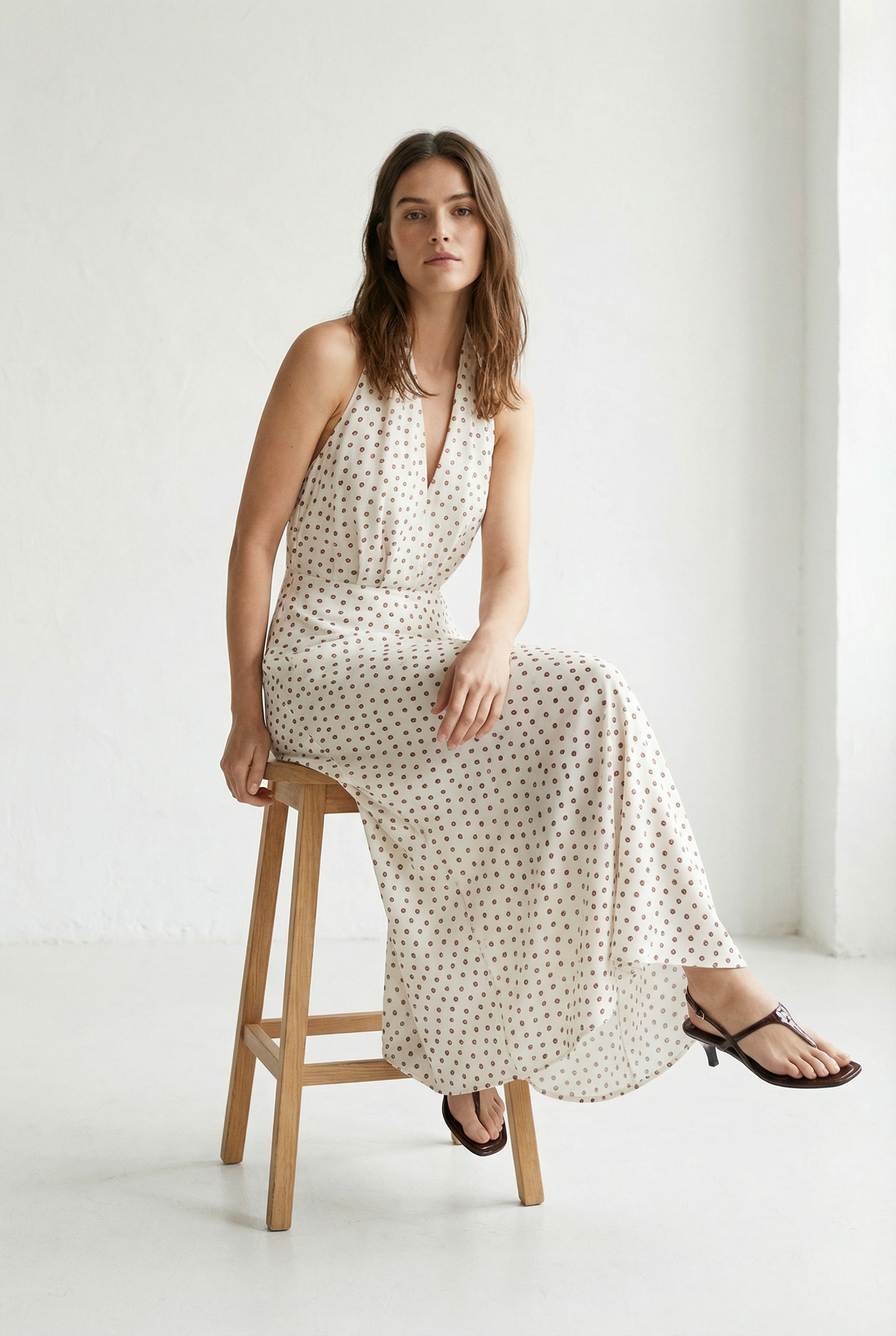 Femme assise dans une position décontractée portant une robe midi imprimée écru à pois, col halter décolleté plongeant.