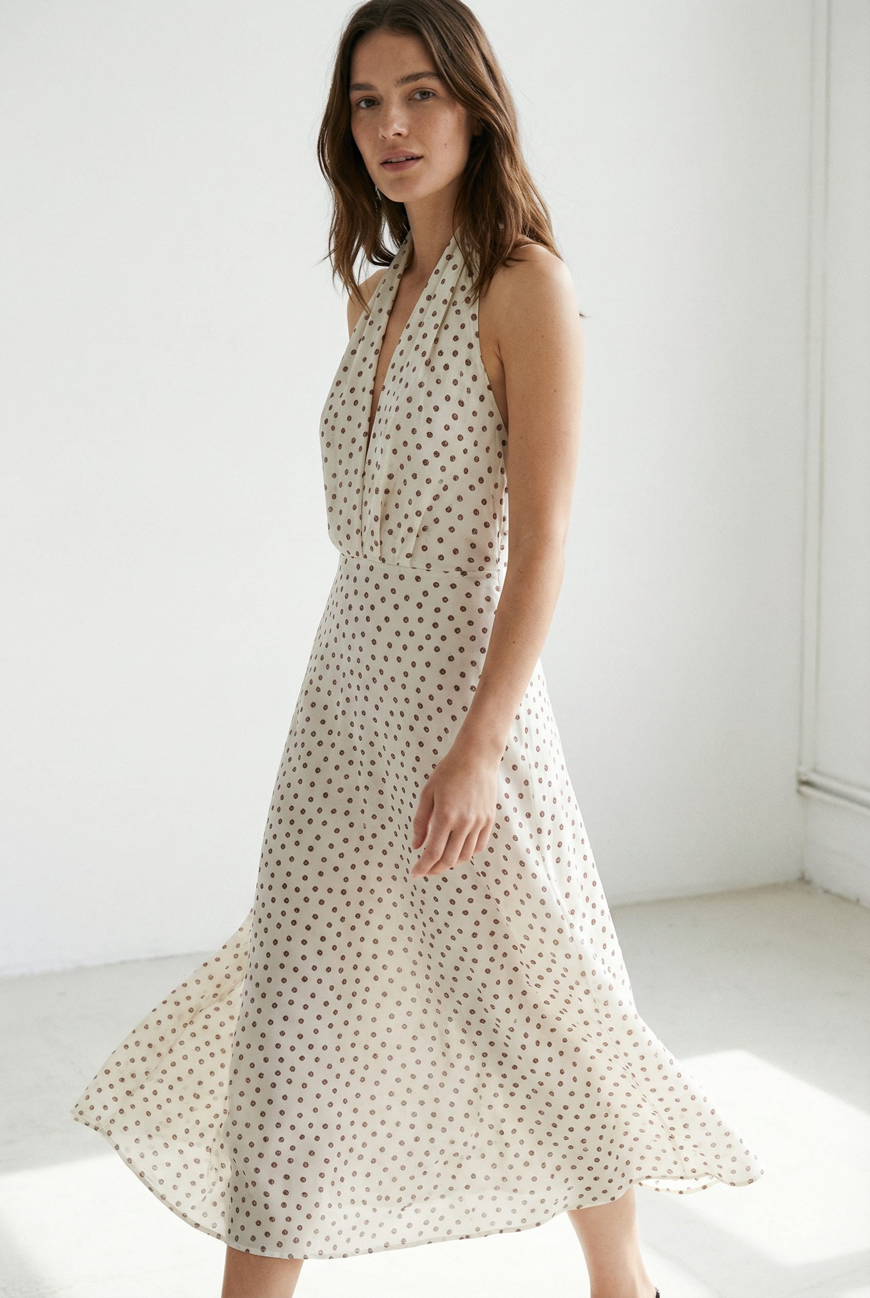 Femme en mouvement dans une robe midi imprimée écru à pois, col halter décolleté plongeant, dans un intérieur lumineux.