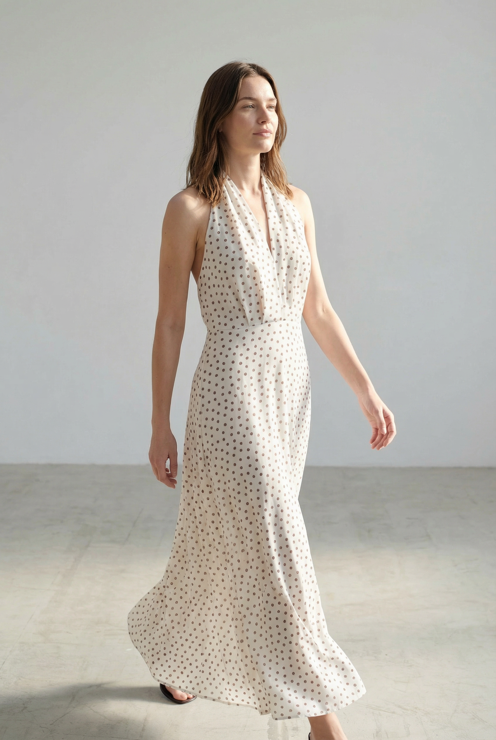 Femme en robe midi imprimée écru à pois, col halter décolleté plongeant, s'avançant avec assurance.