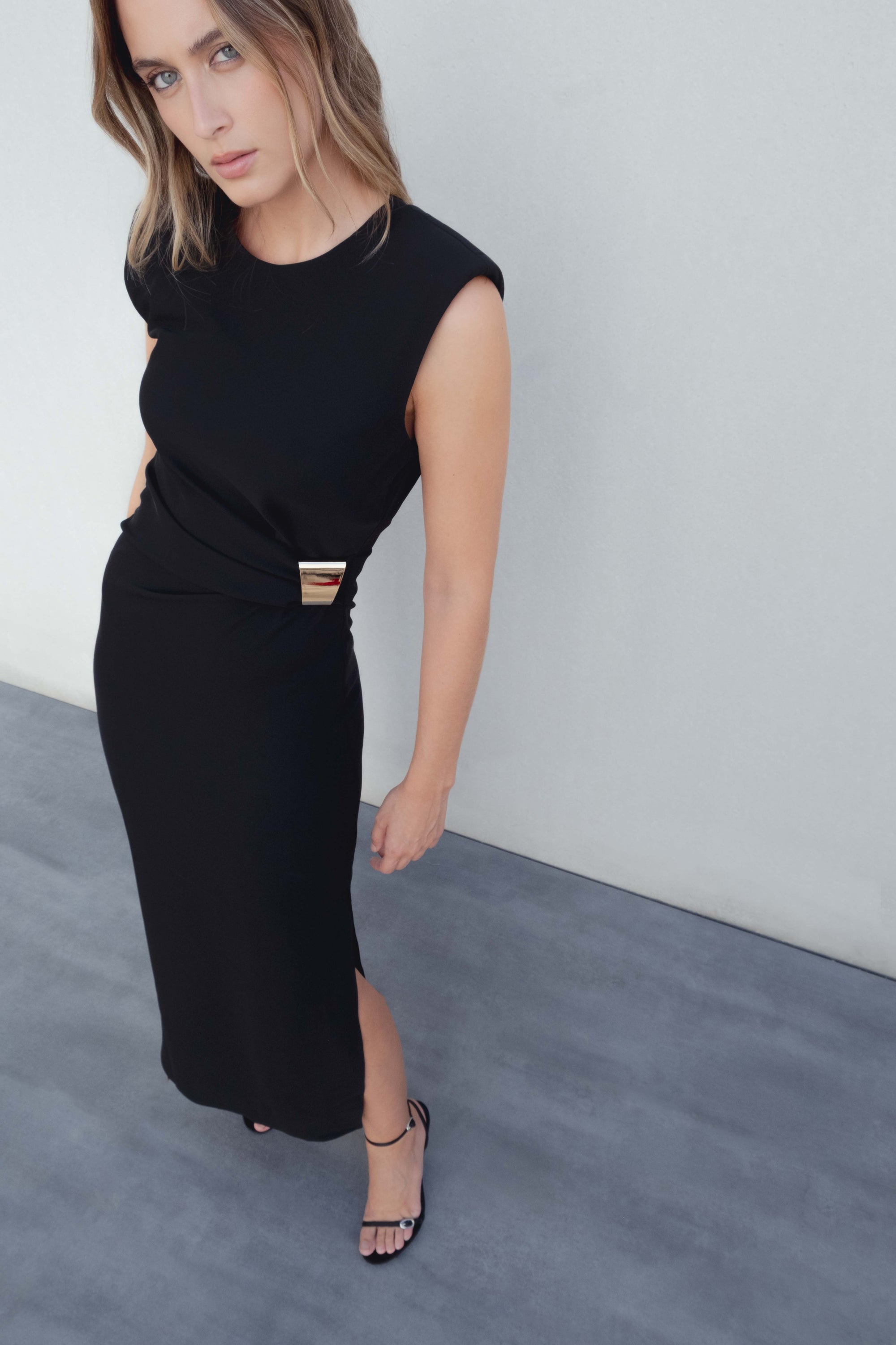 Robe midi noire élégante à applique dorée, modèle porté par un mannequin avec fente latérale et sandales.