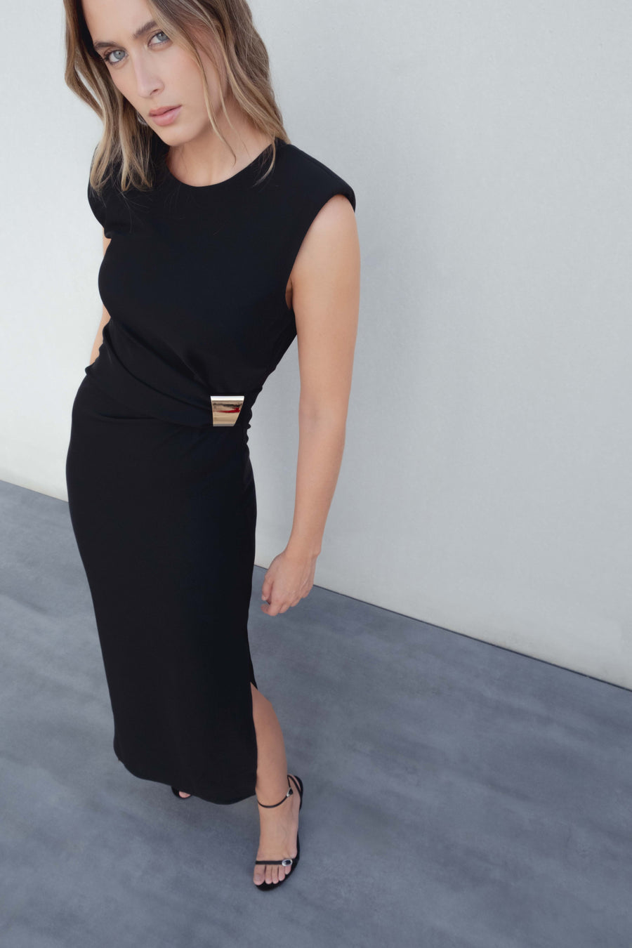 Robe midi noire élégante à applique dorée, modèle porté par un mannequin avec fente latérale et sandales.