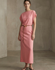Femme élégante en robe midi satinée avec ceinture fine couleur rose, posant dans un intérieur moderne.