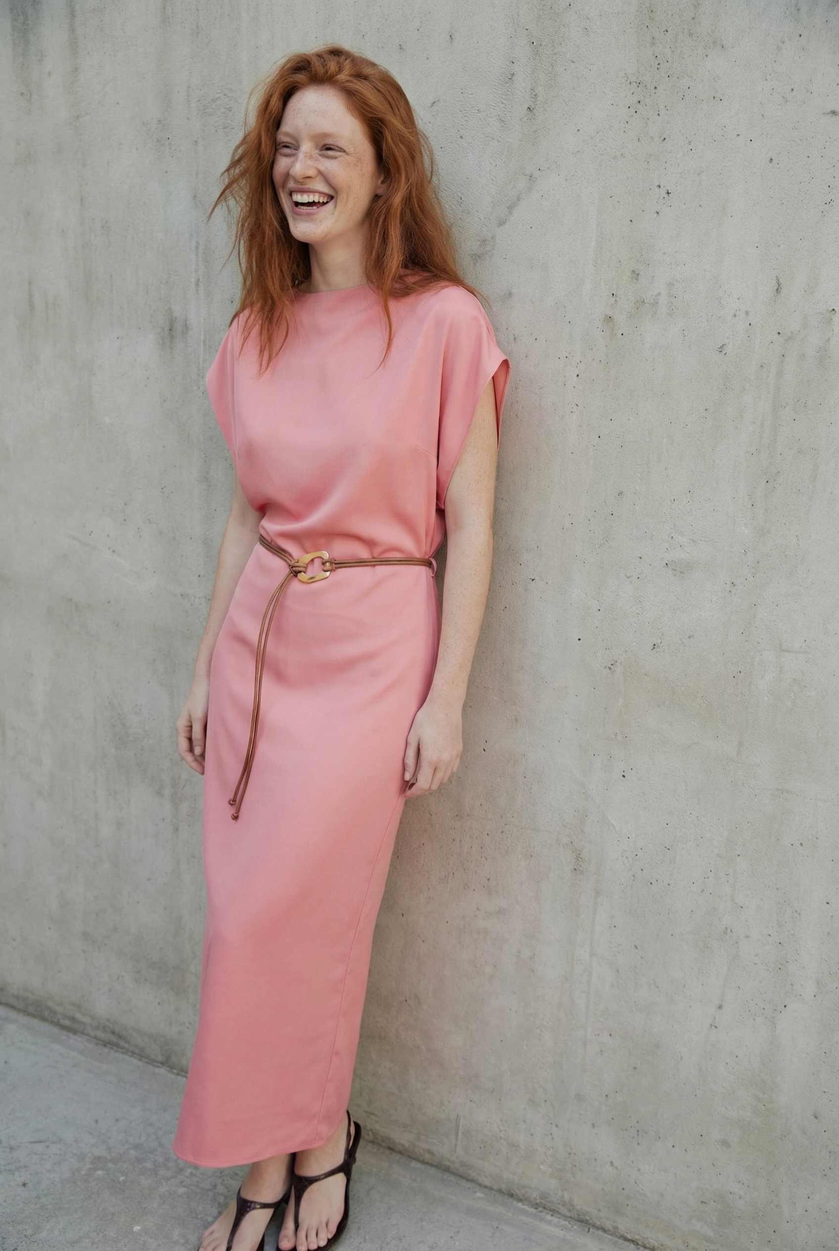 Femme rousse riant joyeusement dans une robe midi satinée avec ceinture fine couleur rose, devant un mur brut.