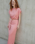 Femme rousse riant joyeusement dans une robe midi satinée avec ceinture fine couleur rose, devant un mur brut.