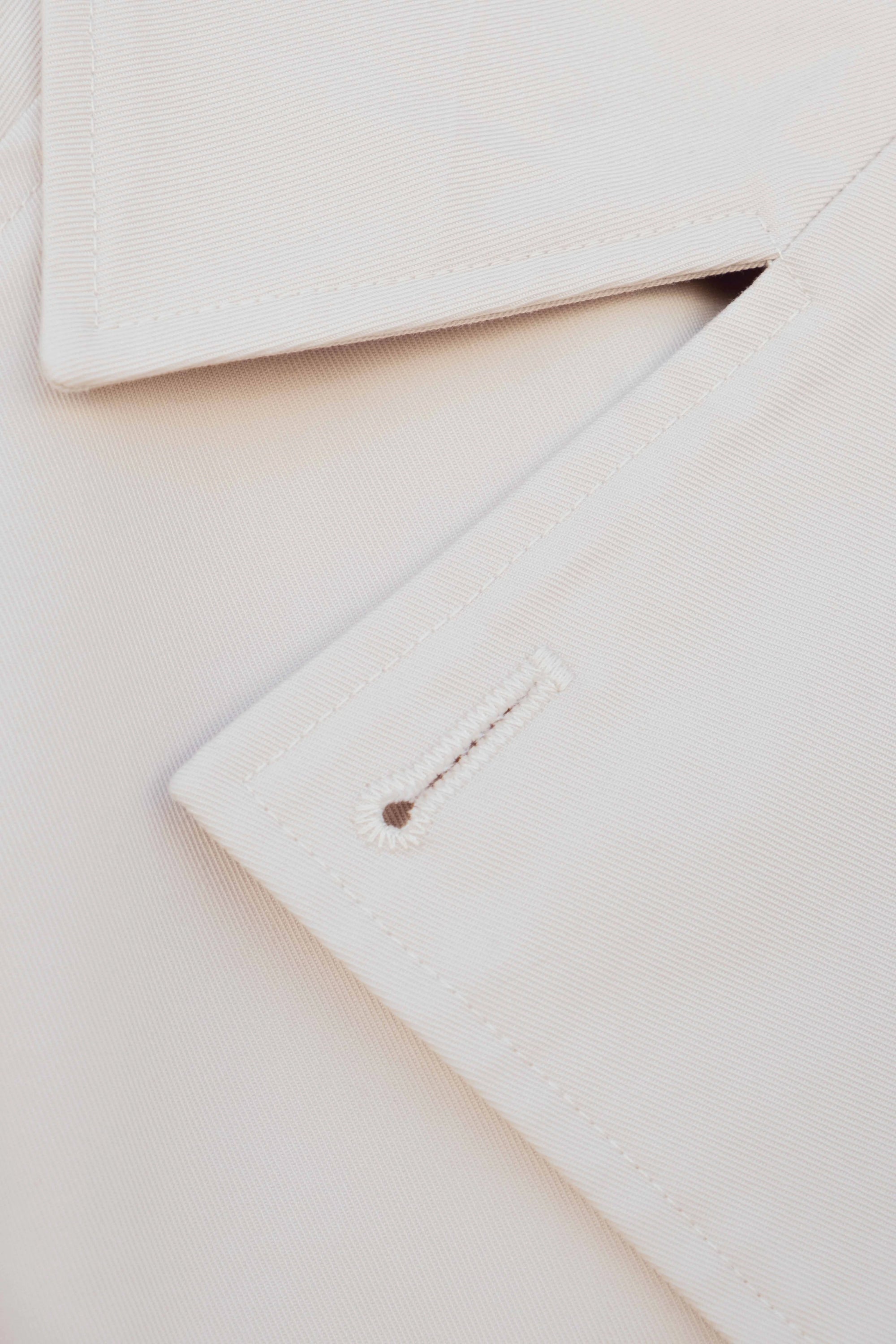 Détail du tissu d'un trench coat court femme beige à double boutonnage, soulignant la qualité et le design élégant de la pièce.