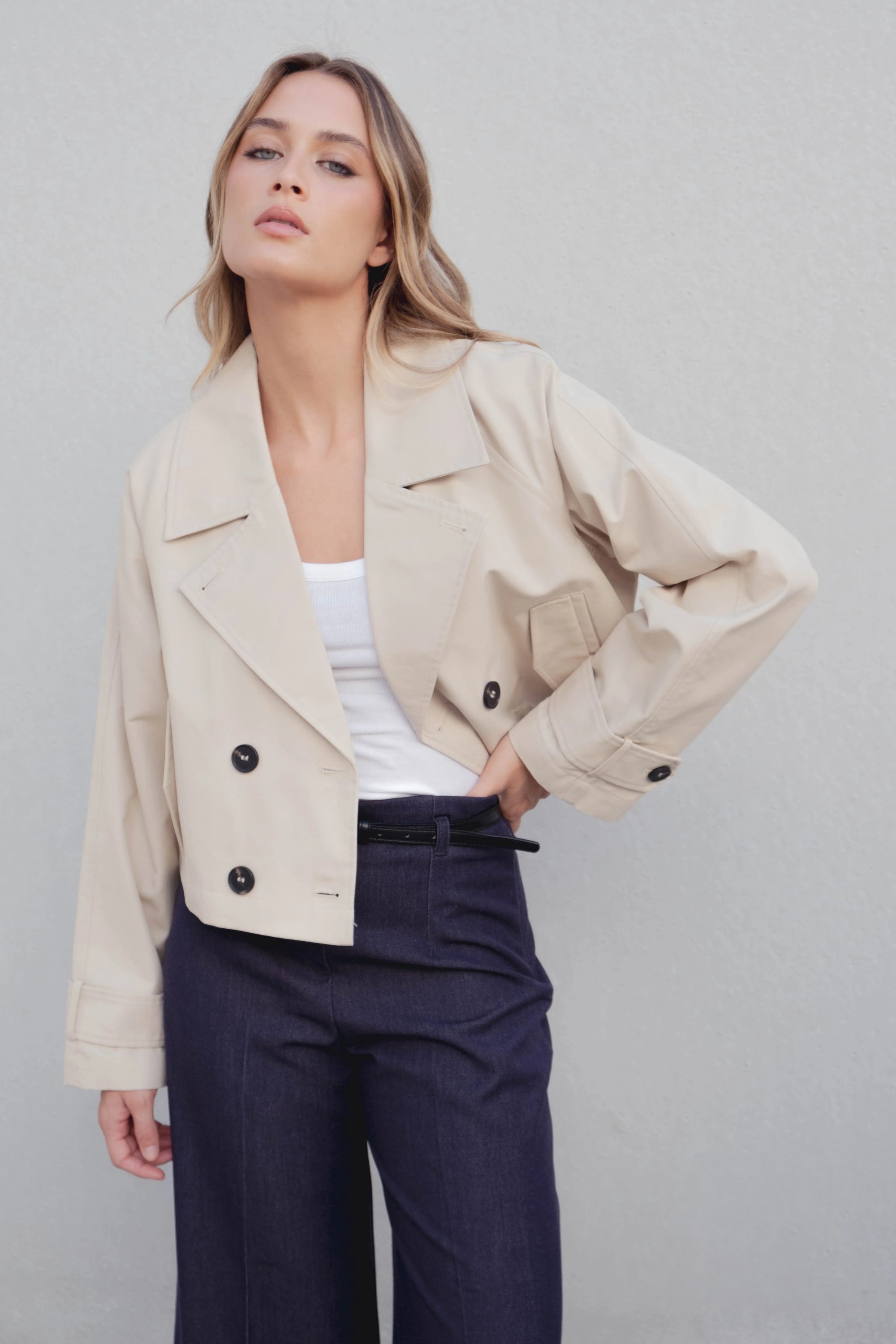 Modèle portant un trench coat court femme beige à double boutonnage, stylé avec un haut blanc et un pantalon sombre, parfait pour un look chic.