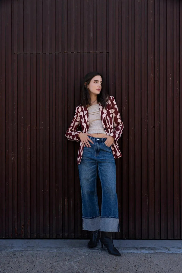 Femme debout avec un blazer imprimé tendance femme chic, en motif graphique, portant un haut beige et un jean, illustrant le look raffiné et moderne du blazer motif femme.