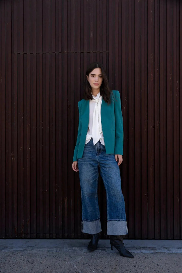 Femme portant un blazer vert ajusté sans col, parfait pour toutes les occasions, associé à une blouse blanche et un jean décontracté. Idéal pour un style chic et contemporain.