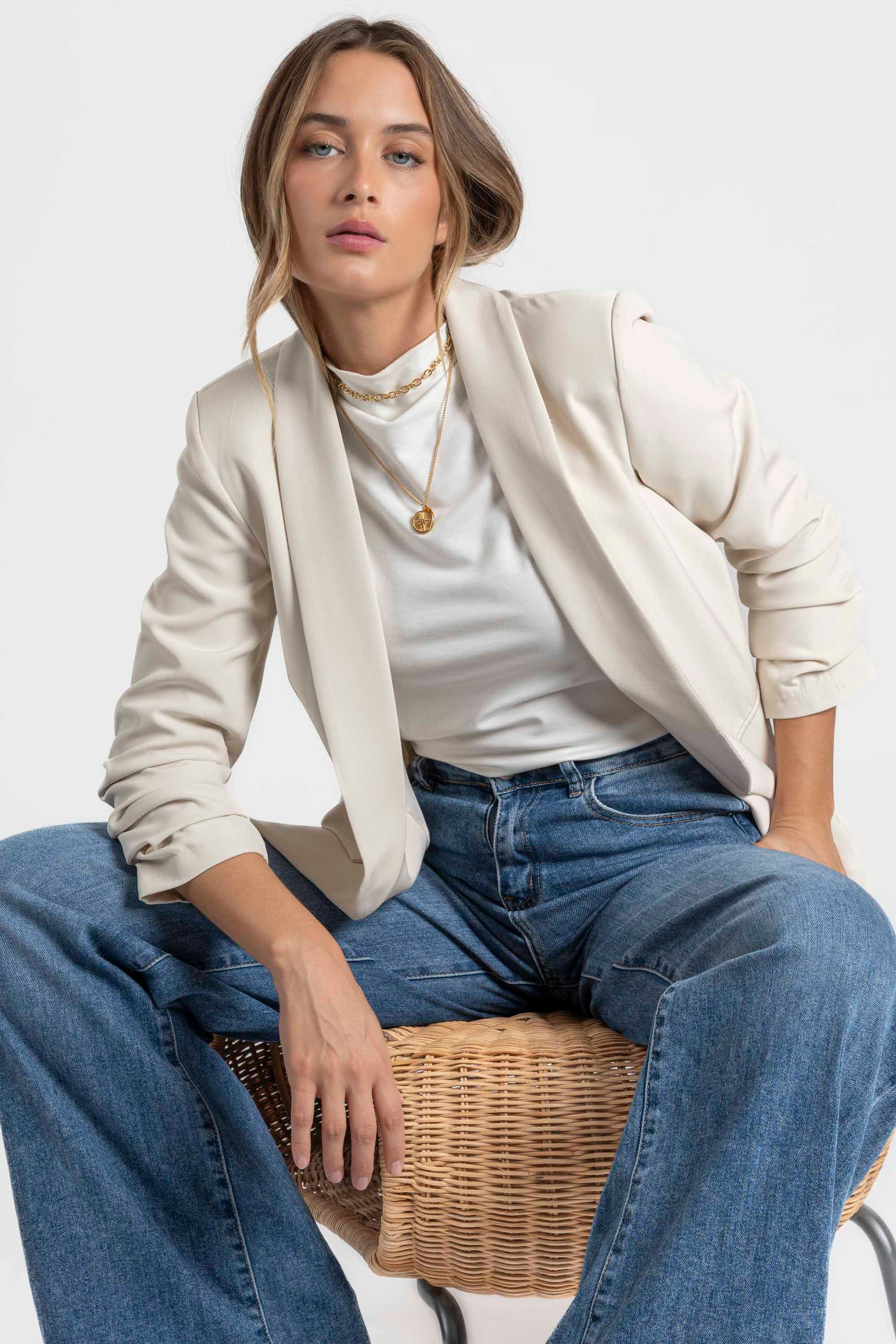 Femme stylisée avec un blazer femme chic crème, se décontractant sur une chaise en osier.