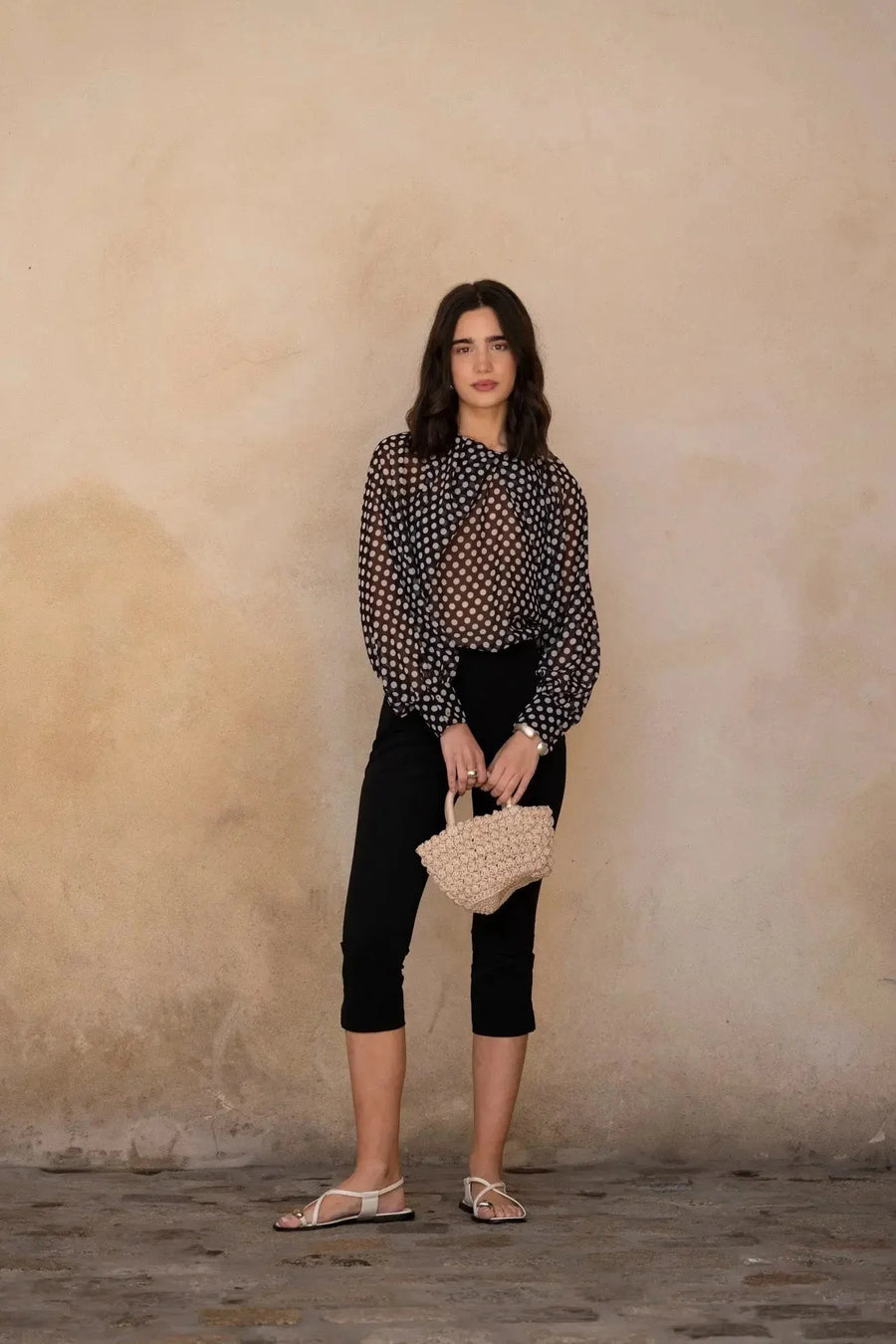 Femme stylée dans un chemisier à pois noire transparente et un pantalon noir, accessoirisé avec un petit sac, un look chic et tendance pour toutes occasions. Chemisier à pois femme.