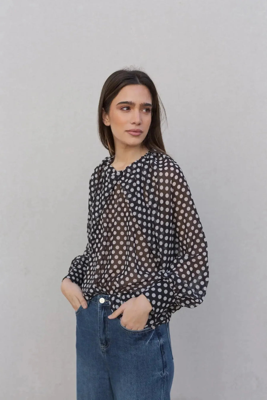 Femme portant un chemisier à pois noire transparente avec des manches bouffantes, stylée avec un jean, parfait pour un look décontracté. Chemisier à pois femme.