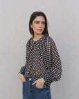 Femme portant un chemisier à pois noire transparente avec des manches bouffantes, stylée avec un jean, parfait pour un look décontracté. Chemisier à pois femme.