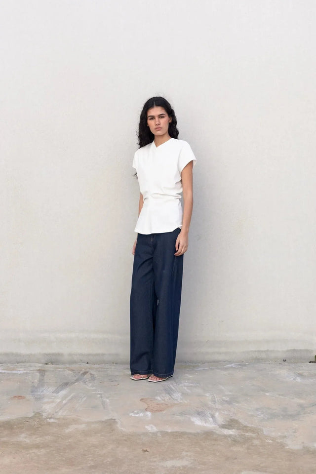 Femme en blouse blanche élégante col V minimaliste, avec un pantalon ample, contre un mur simple. La blouse blanche femme apporte une touche de raffinement à cette tenue.