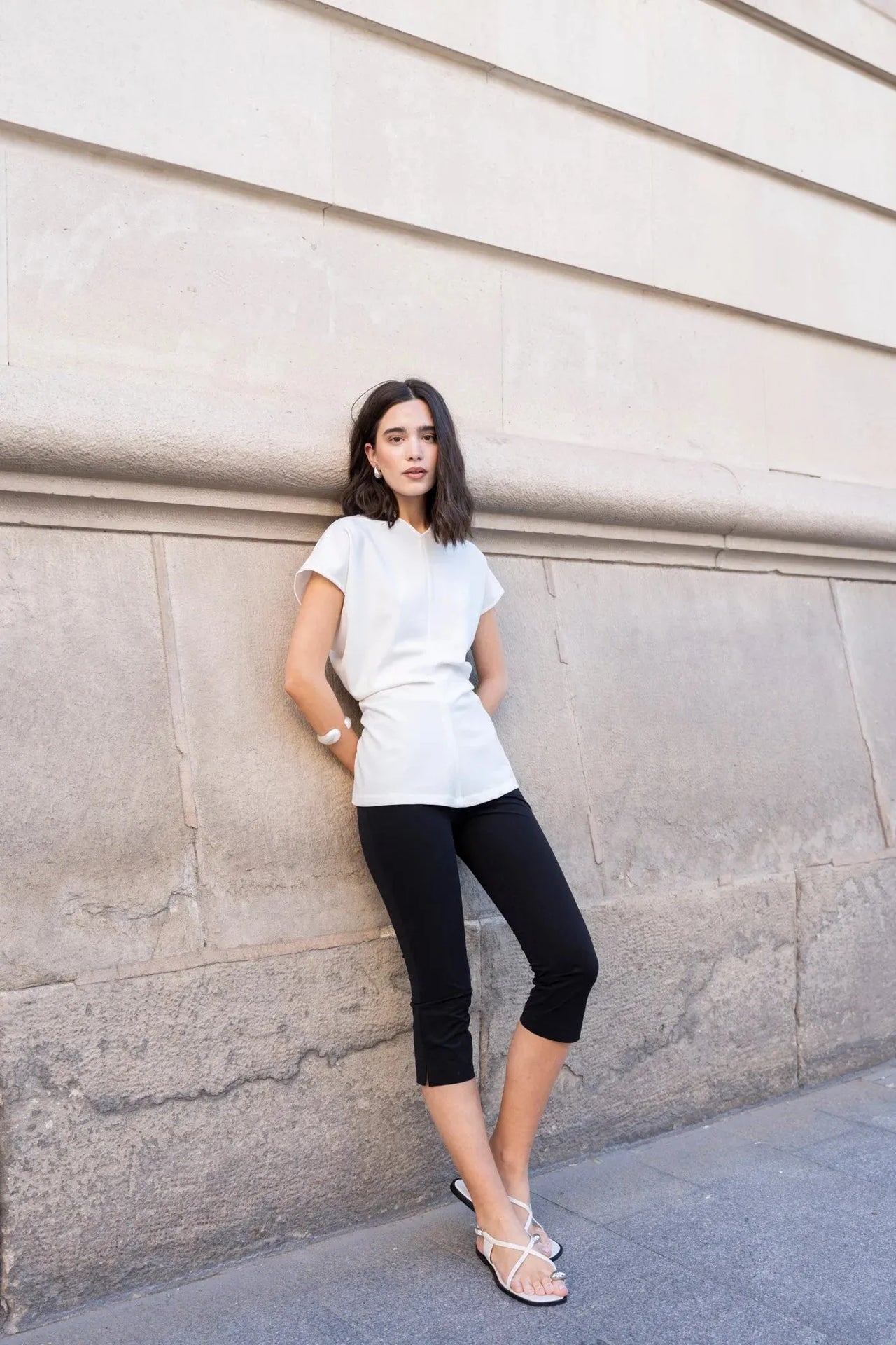 Femme élégante en blouse blanche minimaliste, devant un mur en pierre, associé à un pantalon noir. Parfaite pour un style casual chic, cette blouse blanche femme est à la mode.
