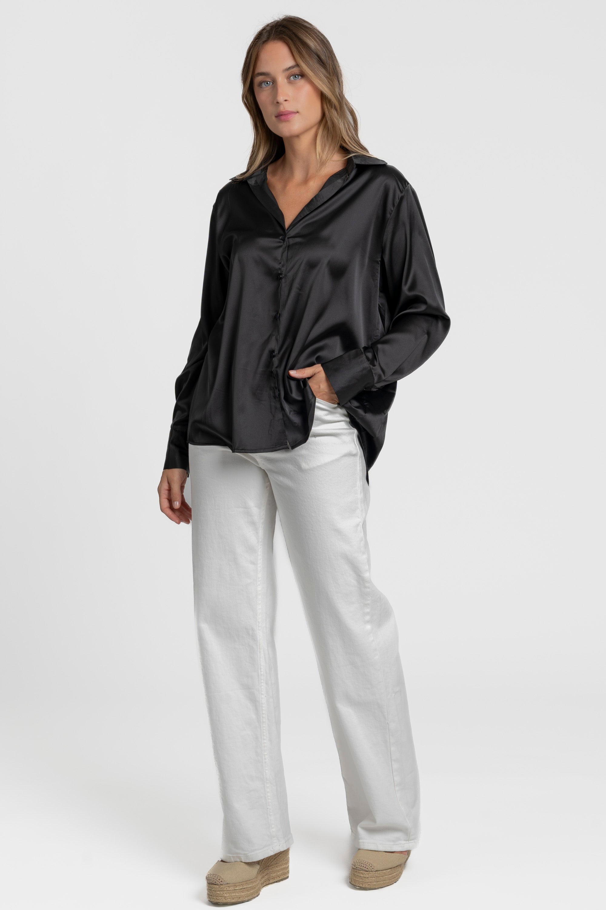 Blouse satinée femme élégante en noir, portée par un modèle, avec une tenue décontractée comprenant un pantalon blanc.