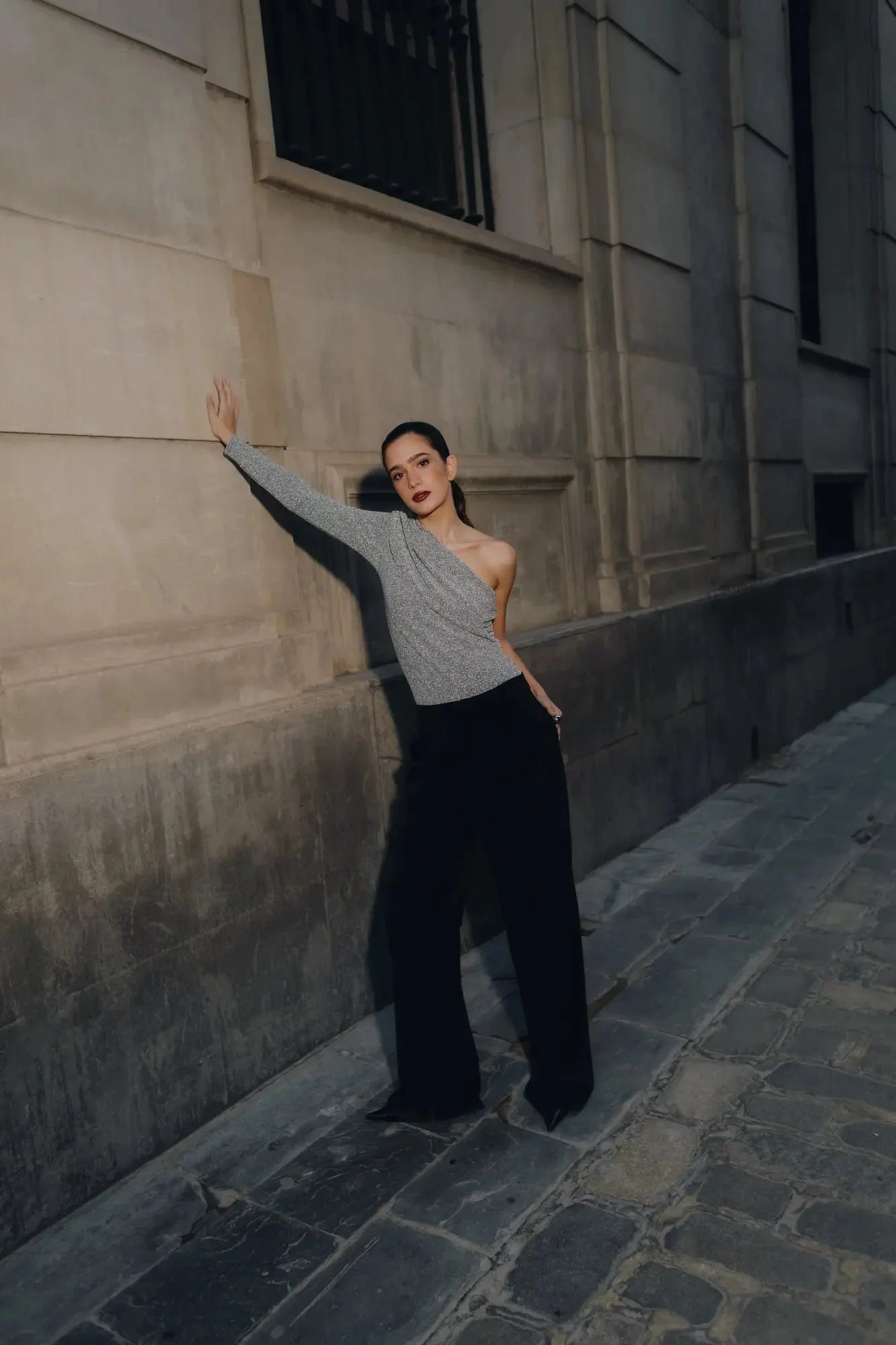 Modèle portant un body asymétrique avec une manche, se tenant contre un mur, illustrant l'élégance unique pour chaque occasion.