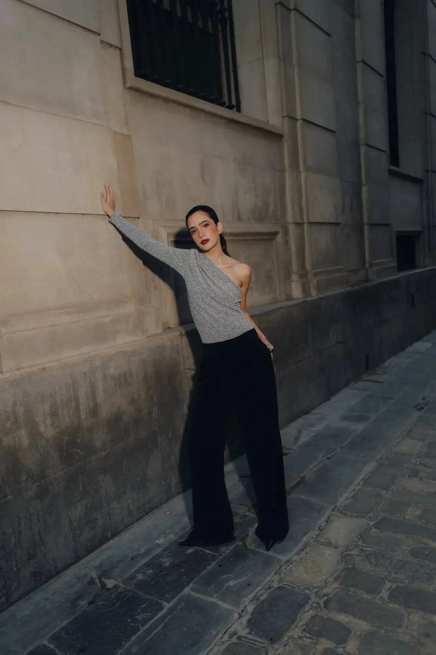 Modèle portant un body asymétrique avec une manche, se tenant contre un mur, illustrant l'élégance unique pour chaque occasion.