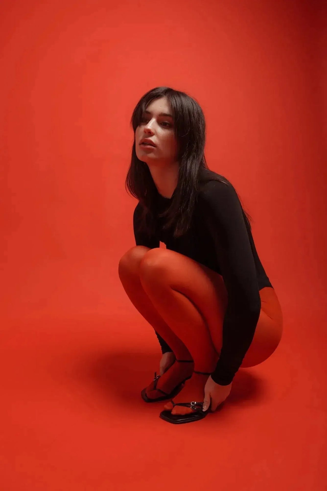 Body Drapé Noir Style et Confort Réunis - Femme en position accroupie, portant un body manche longue femme, sur fond rouge, illustrant la polyvalence et le confort.