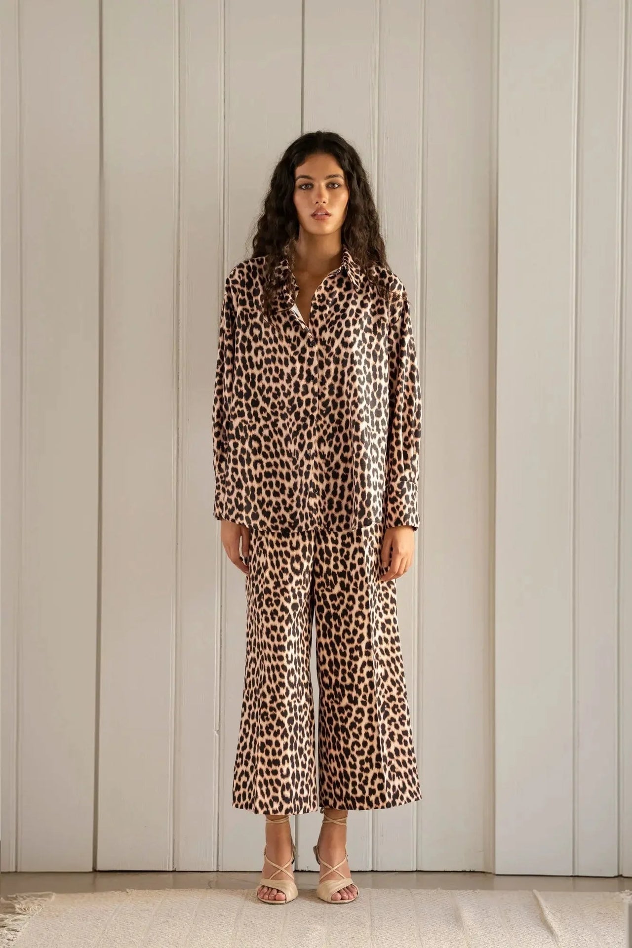 Femme debout dans un décor sobre, portant une chemise à imprimé léopard à manches longues et un pantalon assorti. Chemise longue leopard femme.