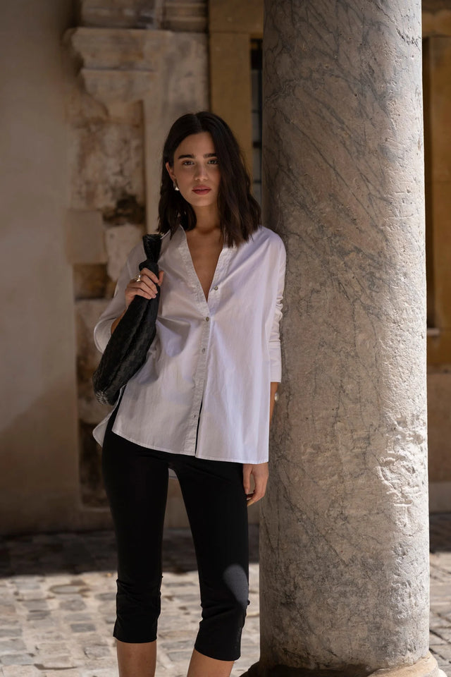 Chemise Femme Blanche Ample en popeline, portée par un modèle devant un pilier dans un cadre urbain élégant.