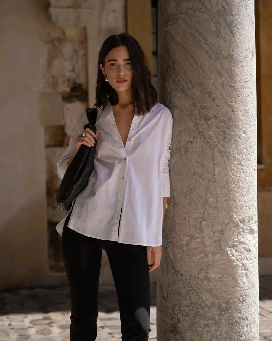 Chemise Femme Blanche Ample en popeline, portée par un modèle devant un pilier dans un cadre urbain élégant.