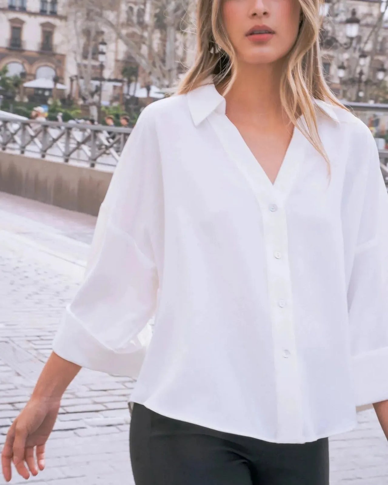 Une chemise fluide blanche à manches larges et dos fendu, portée par une femme marchant sur une promenade, mettant en avant le style décontracté de ce chemisier ample femme.