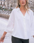 Une chemise fluide blanche à manches larges et dos fendu, portée par une femme marchant sur une promenade, mettant en avant le style décontracté de ce chemisier ample femme.