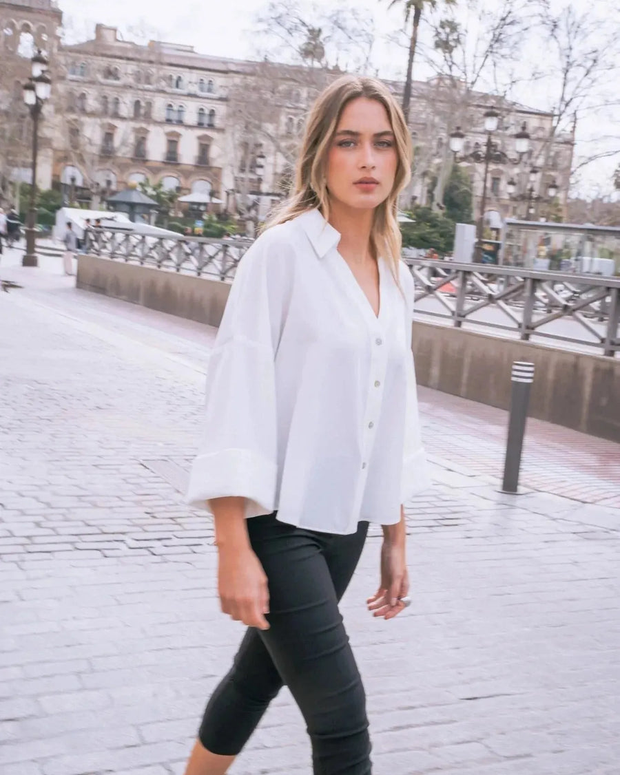 Une chemise fluide blanche à manches larges et dos fendu, présentée sur une femme marchant, soulignant l'élégance décontractée de ce chemisier ample femme.