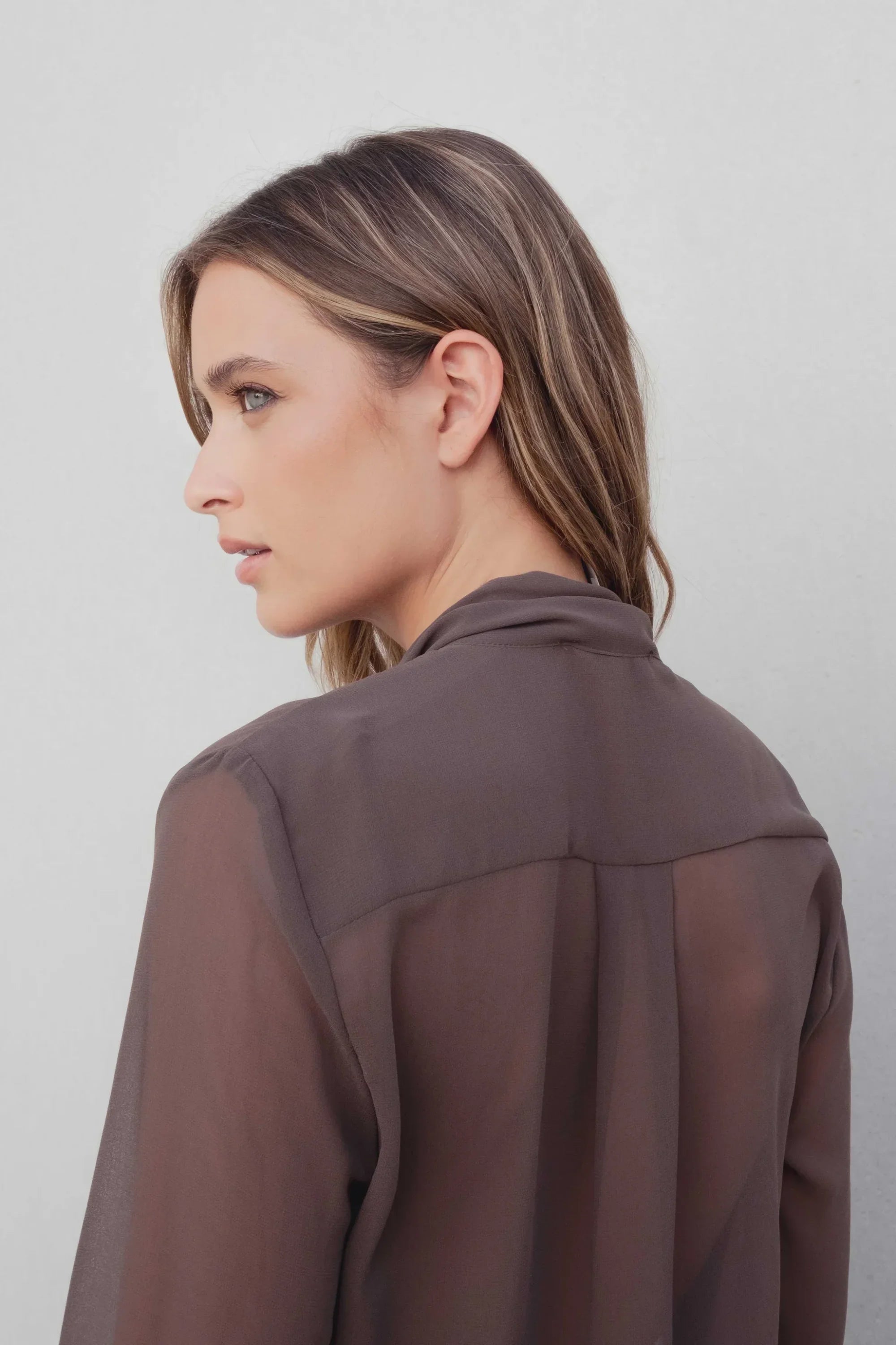 Vue latérale d'une femme portant une chemise col lavallière fluide marron transparent, montrant le col et la coupe sophistiquée de la chemise col lavallière.
