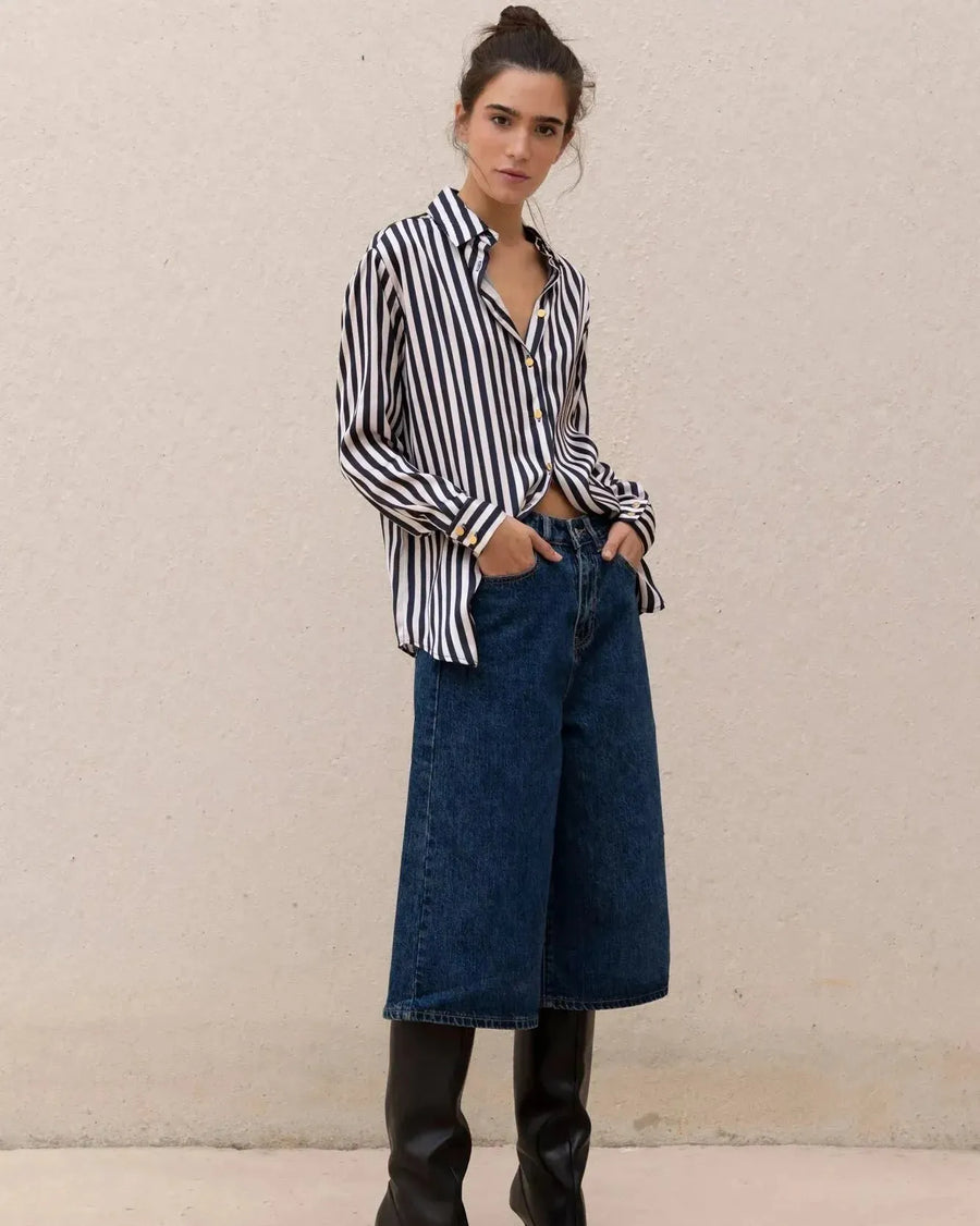 Femme debout en chemise rayée bleu marine et blanc devant un mur neutre, combinée avec un jean en denim et des bottes élégantes. Un choix parfait pour un look chic avec la chemise rayée femme.