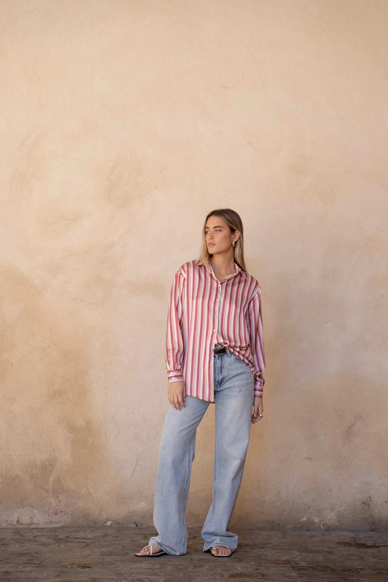 Chemisier satin femme chic à rayures verticales, couleur rose et beige, présenté avec un style décontracté, mannequin se tenant debout dans un ambiance minimaliste.
