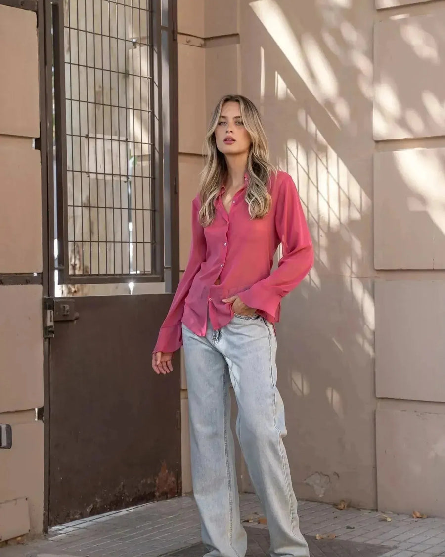 Femme posant avec confiance en chemise transparente rose à manches longues pour femme, accompagnée de jeans, pour une tenue mode et sans effort. Restez élégante avec la chemise transparente femme.