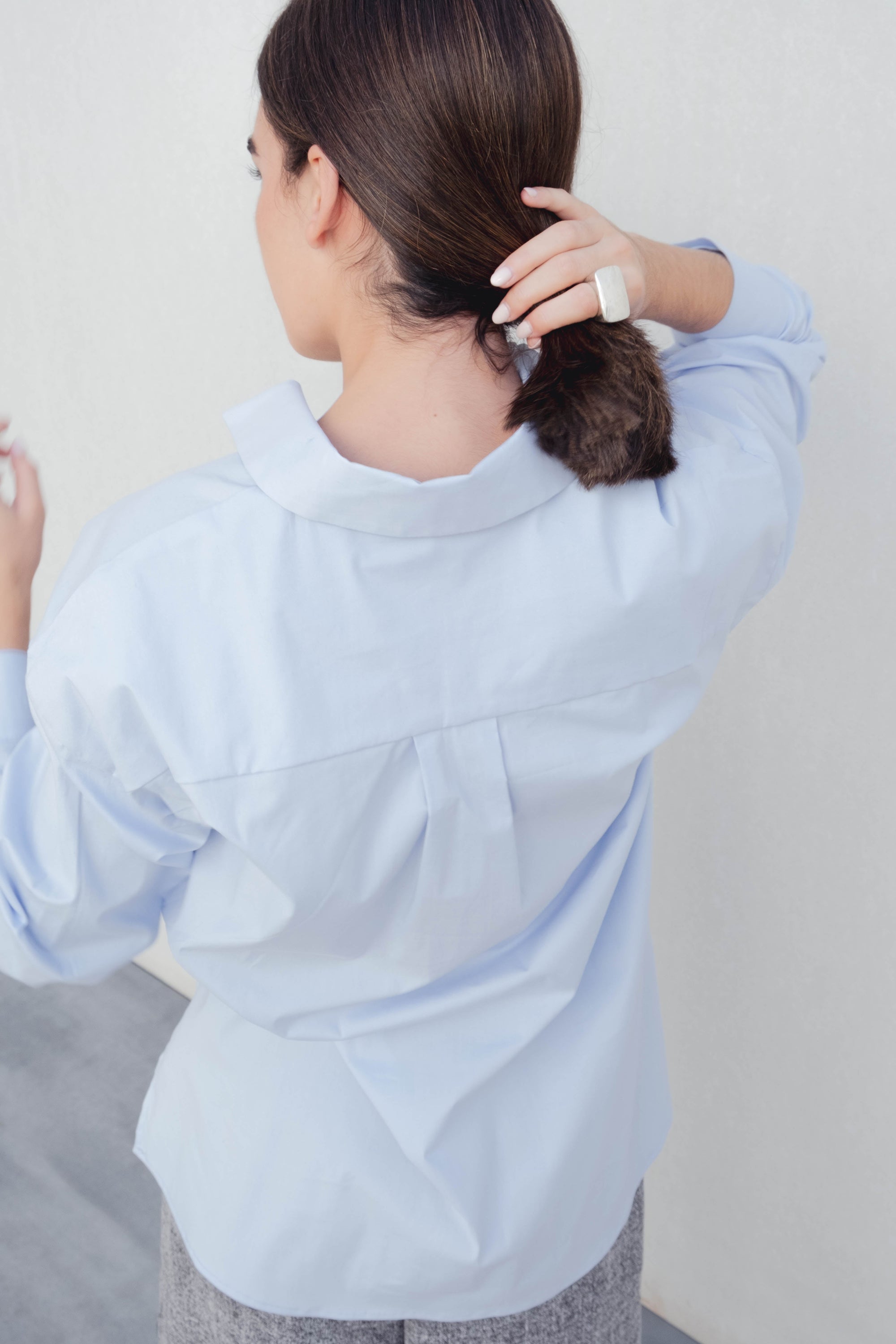 Vue arrière d'une femme ajustant ses cheveux tout en portant une chemise oversize popeline bleu ciel, mettant en avant le style chic et confortable de cette chemise oversize femme.