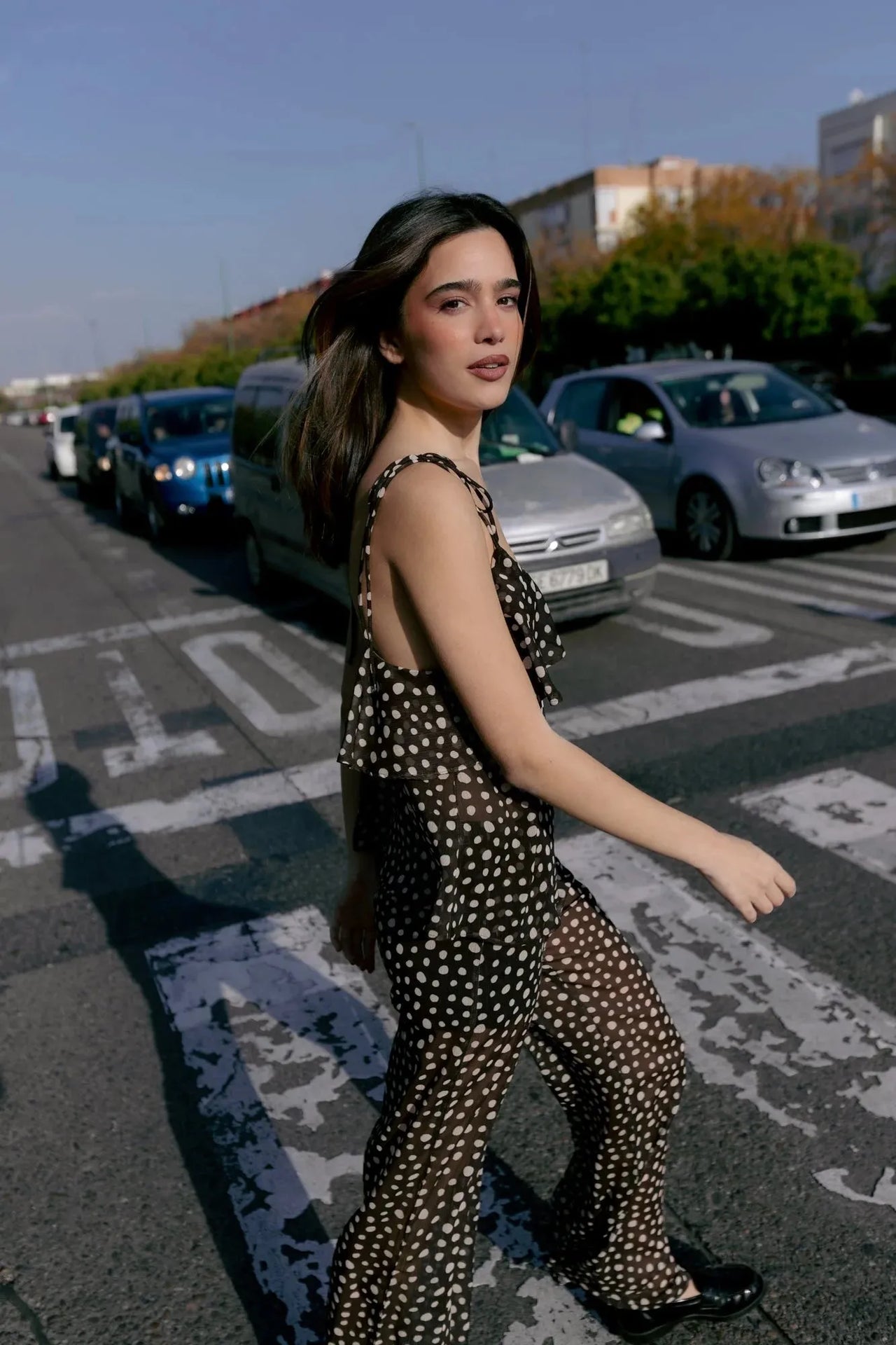 Femme marchant dans la rue avec une combinaison fluide à pois blancs et volants, un choix élégant et décontracté pour l'été, accompagnée d'un chemisier voile transparent femme.